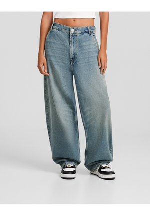 Personne portant un jean large bleu clair taille haute et des baskets noir et blanc, debout sur un fond gris uni.