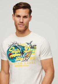 Camiseta de manga corta blanca con un diseño gráfico colorido que presenta olas, una tabla de surf y texto. Material de algodón con una textura suave.