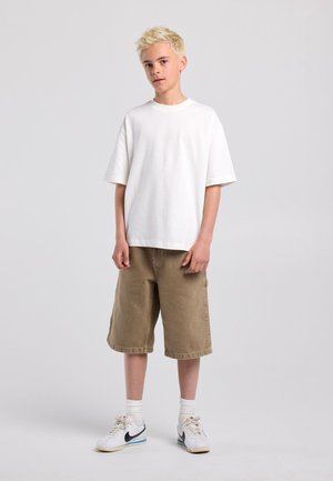 Blonder Teenager-Junge steht, trägt ein locker sitzendes weißes Kurzarmhemd, knielange braune Shorts, weiße Socken und weiße Nike-Sneaker.