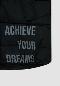 Chaqueta negra acolchada con gran texto gris "LOGRA TUS SUEÑOS" en el frente. Tejido liso y brillante con diseño acolchado.