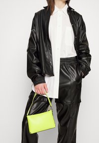 Svart fusk läderjacka och byxor med en vit skjorta under. Hand som håller en liten neongrön handväska. Slät textur och minimalism.