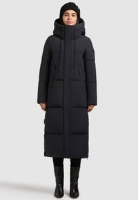Lungo cappotto piumino nero con cappuccio, con sezioni quadrate trapuntate, corde regolabili e tasche frontali. Indossato sopra pantaloni neri e stivali.