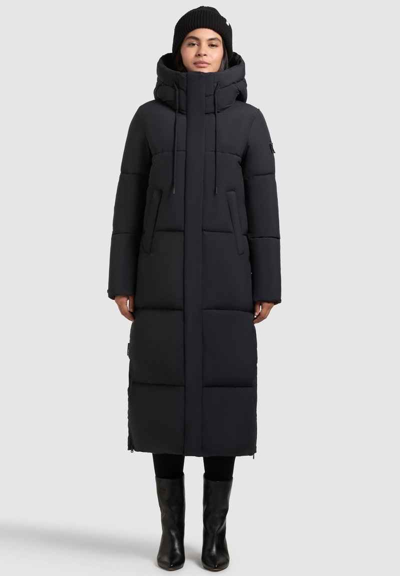 Lungo cappotto piumino nero con cappuccio, con sezioni quadrate trapuntate, corde regolabili e tasche frontali. Indossato sopra pantaloni neri e stivali.