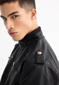 Chaqueta de piel negra con un cuello alto, bolsillos con cremallera y detalles de hardware en plateado. Textura suave y diseño ajustado.