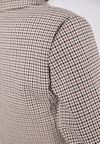 Tissu à motif pied-de-poule brun et blanc avec des accents noirs, présentant une surface texturée et une coupe ajustée, vu de côté.