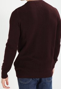 Burgundy tröja med en texturerad herringbone-mönster, rundad hals, och ribbade ärmslut och kant, kombinerad med blå jeans.