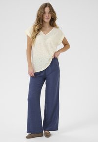 Top giallo chiaro, strutturato, con scollatura a V e maniche corte, abbinato a pantaloni larghi blu scuro. Scarpe marroni completano l'outfit.
