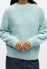 Jersey de punto azul claro con cuello redondo, puños y dobladillo acanalados, con un patrón texturizado y ajuste relajado. Las manos están entrelazadas al frente.