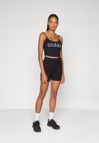 Svart avkortet tanktop med "GUESS"-logo, kombinert med svarte sykkelshorts. Modellen har på seg svarte sneakers og hvite sokker. Nøytral bakgrunn.