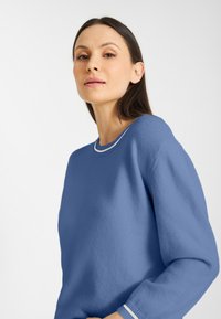 Vrouw met donker haar in een blauwe trui met een wit-omrandde kraag en manchetten, die iets omhoog kijkt tegen een effen witte achtergrond.