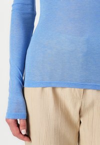 Helles blaues Langarmoberteil aus dünnem, glattem Stoff, kombiniert mit zerknitterten beigen Hosen. Hat einen runden Ausschnitt und schmale Ärmel.