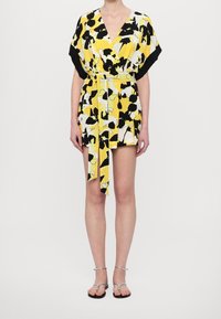 Femme portant une robe portefeuille à fleurs jaune, noire et blanche avec une ceinture nouée et des sandales plates argentées à brides, debout devant un fond uni.