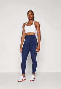 Μπλε ναυτικού ψηλόμεσο κολάν με λουλουδάτο πάνελ διακόσμησης, συνδυασμένο με λευκό αθλητικό σουτιέν. Το μοντέλο φοράει λευκά και πορτοκαλί αθλητικά παπούτσια.