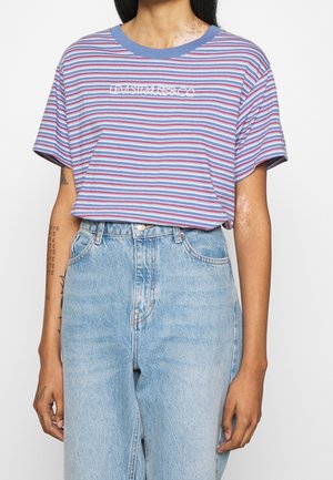 Personen har højttaljede lyseblå jeans på og en blå, rød og hvid stribet t-shirt med teksten "LEVI'S P4US8 & CO" på brystet.