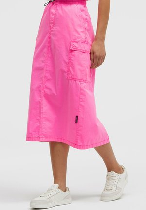 Frau trägt einen leuchtend pinken Midirock mit seitlicher Cargo-Tasche und weißen Sneakers, steht vor einem schlichten Hintergrund.