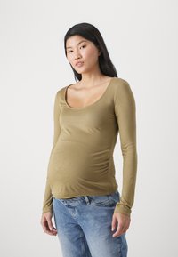 Top de maternidad de manga larga en verde oliva, fabricado en tejido elástico, con escote redondeado y costuras laterales fruncidas para mayor comodidad. Combinado con unos jeans azul claro.
