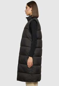Gilet sans manches noir en doudoune avec un col haut, doté d'un design matelassé et d'une fermeture éclair sur le côté. Tissu lisse avec une texture légère et gonflée.