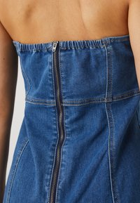 Denimové šaty s přiléhavým siluetou, s nařaseným horním okrajem, viditelným zipovým zapínáním a kontrastním prošíváním v středně modré barvě.