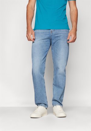 Homme portant un jean droit bleu clair, des baskets blanches et un t-shirt bleu vif, debout devant un fond uni.