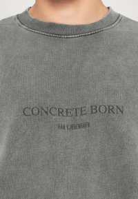 Arti šviesiai pilka marškinėlių su apvaliu kaklu detalė, ant krūtinės spausdinta užrašas "CONCRETE BORN" ir mažesnis tekstas "HAY K. COPENHAGEN".