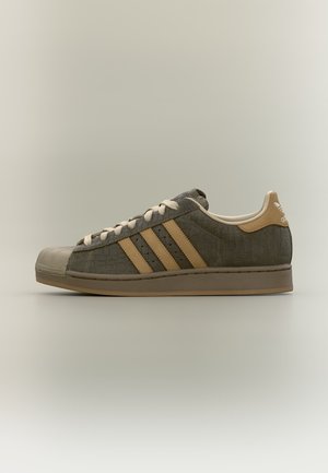 Basket basse Adidas avec empeigne marron foncé texturée, bandes beige, lacets, languette au talon et semelle en caoutchouc sur fond neutre.