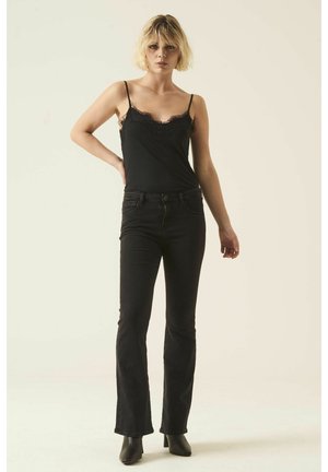 Schwarzes Camisole mit Spitzenbesatz, kombiniert mit figurbetonten schwarzen Schlagjeans; beide haben eine glatte Textur und eine schlanke Silhouette. Das Model steht in einem neutralen Umfeld.