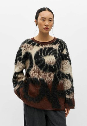 Mulher de cabelo escuro veste um camisola felpuda castanha, preta e creme com padrão e calças cinzentas escuras, em pé contra um fundo branco simples.