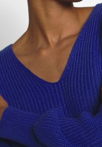 Blå stickad tröja med V-ringning och texturerade mönster. Tyget verkar mjukt med synlig ribbdetalj.