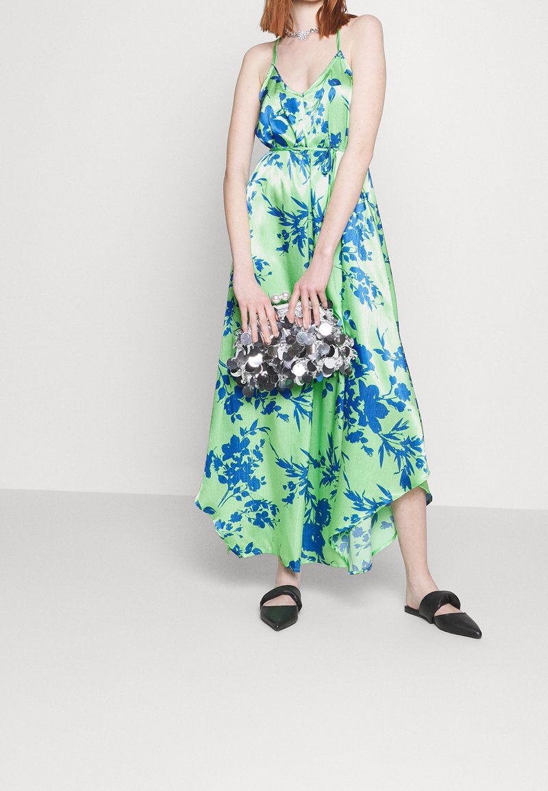 Femme en robe sans manches fleurie verte et bleue tenant un sac à main argenté à sequins, portant des ballerines noires pointues, debout sur un fond uni.