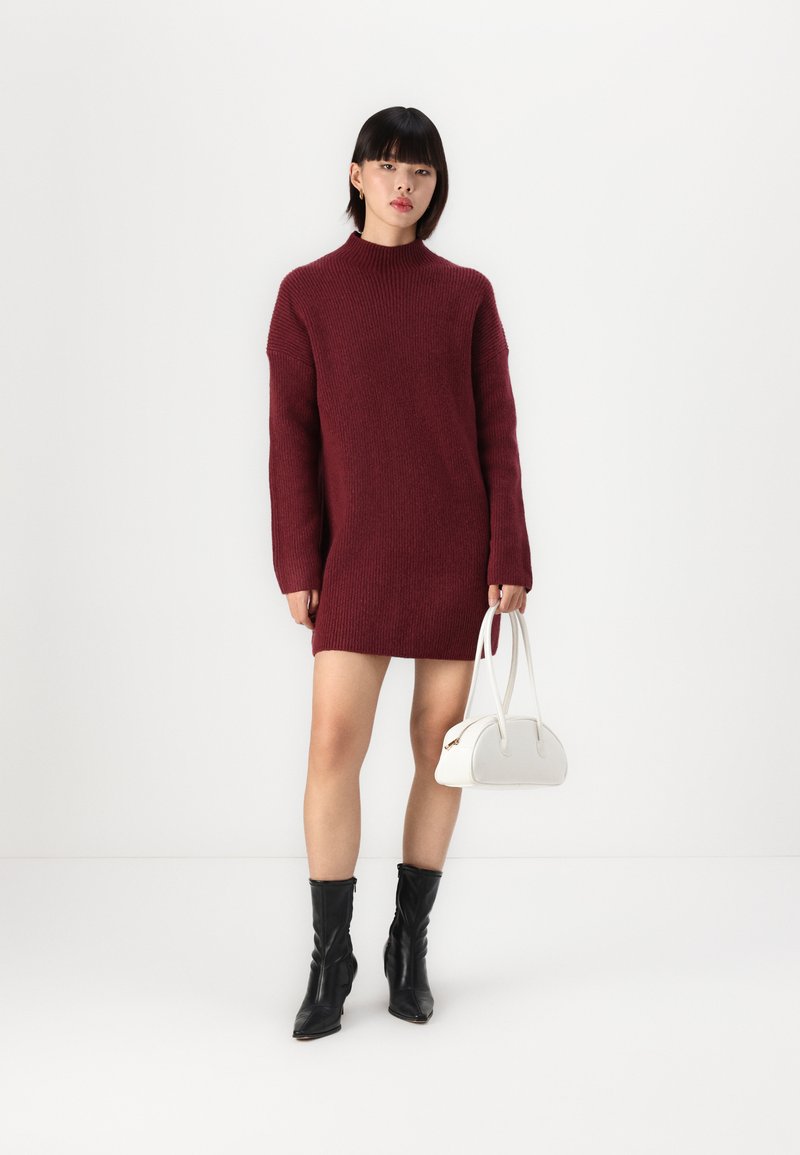 Kuscheliges, kirschrotes Strickkleid mit hohem Kragen, langen Ärmeln und einem lockeren Schnitt. Kombiniert mit schwarzen Ankle-Boots und einer weißen Handtasche.