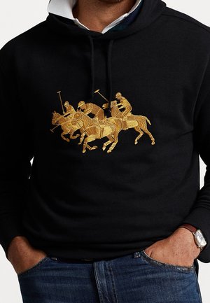 Sweat à capuche noir avec broderie dorée de trois joueurs de polo à cheval tenant des maillets, porté sur une chemise blanche à col.
