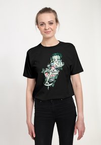 Sort t-shirt med et farverigt grafik af en karakter med grønt hår og lyserøde klæder, omgivet af blomstrelementer og et banner.