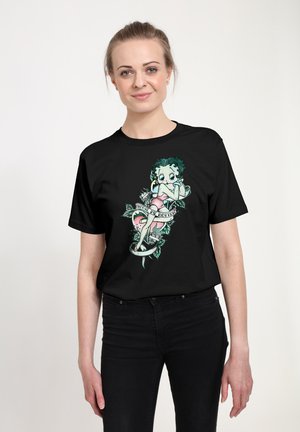 Sort t-shirt med et farverigt grafik af en karakter med grønt hår og lyserøde klæder, omgivet af blomstrelementer og et banner.