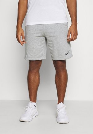 Hombre que lleva zapatillas blancas, calcetines blancos, pantalones cortos grises de Nike y una camiseta blanca, de pie contra un fondo liso.