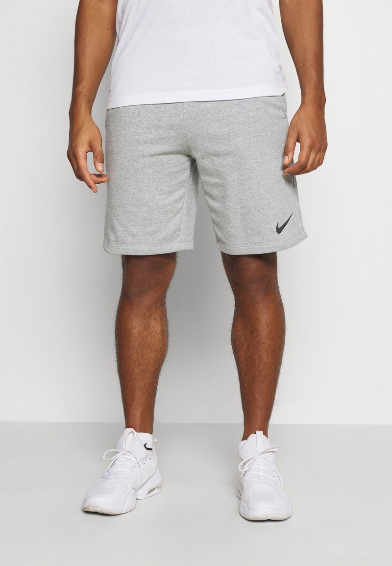 Man som bär vita sneakers, vita strumpor, grå Nike-shorts och en vit t-shirt står mot en enkel bakgrund.