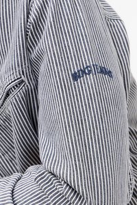 Prugasta traper košulja s tamnoplavim i bijelim vertikalnim prugama. Sadrži izvezeni logo "BDG TEAM" u mornarskoj plavoj boji na gornjoj ruci.