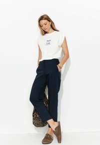 T-shirt blanc à manches courtes avec un texte graphique bleu, associé à un pantalon tailleur bleu marine. Les accessoires comprennent un sac en imprimé léopard et des mules marron.