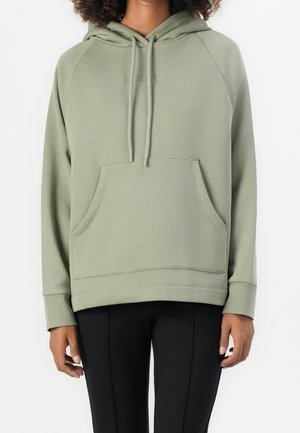 Personne portant un sweat à capuche vert clair avec une poche avant et un pantalon noir, debout devant un fond blanc.