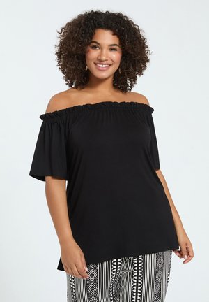 Smilende kvinde med krøllet hår iført en sort skulderfri top og sort-hvide mønstrede bukser, stående foran en ensfarvet baggrund.