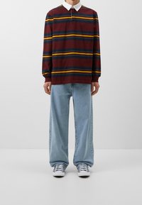 Bordeauxrotes Langarmpolo mit horizontalen Streifen in Blau, Gelb und Grün; kombiniert mit lockeren hellblauen Jeans und grauen Turnschuhen.