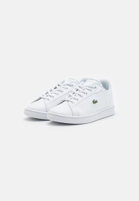 Hvite lave sneakers laget av syntetisk materiale. Funksjoner inkluderer flate lisser, rund tå og en liten grønn krokodillelogo på siden.