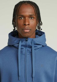 Hoodie zippé bleu en tissu doux, avec un col montant, des cordons de serrage et un logo discret sur la poitrine. Texture lisse et coupe ajustée.