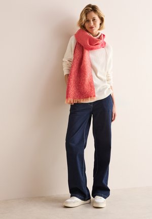 Sciarpa a motivo rosso e rosa appoggiata su un maglione bianco a maglia, abbinata a jeans a gamba larga blu scuro e sneakers bianche. Sfondo semplice.