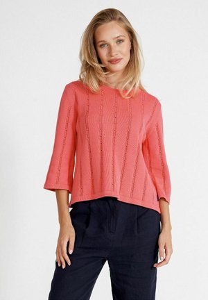 Korallenfarbener Strickpullover mit ¾-Ärmeln, der vertikale Rippenmuster und offene Maschenverzierungen aufweist, kombiniert mit dunkeln marineblauen Hosen.
