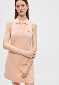 Femme portant une robe polo sans manches couleur pêche avec un col et des boutons, tenant son bras gauche avec sa main droite devant un fond uni.