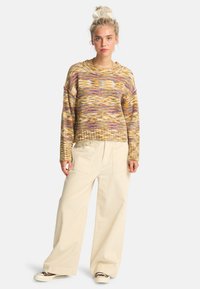 Sweter z dzianiny w mieszanych kolorach żółtym, purpurowym i brązowym. Ma luźny krój i ściągacz u dołu. Połączony z szerokimi beżowymi spodniami.