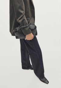 Zwarte geweven handtas met een textuurafwerking en een gevlochten hengsel, vastgehouden in een hand naast een wijde donkere denim broek en een op maat gemaakte grijze blazer.