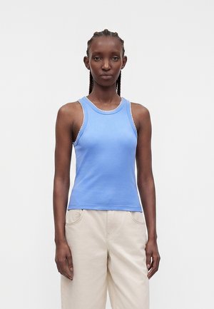 Woman with braided hair wearing a sleeveless light blue top and beige pants, standing against a plain light background.