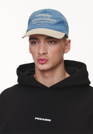 Blå caps med beige skygge, med hvit tekst: "idrettsforening alltid i endring for bedre aldri for sent." Bært med en svart hettegenser.