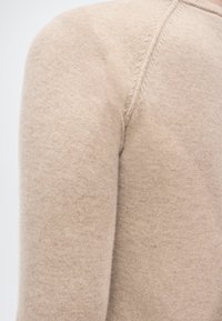 Κοντινό πλάνο μανικιού μπεζ πλεκτού πουλόβερ που δείχνει απαλό, λεπτό μάλλινο ύφασμα με ορατές ραφές στη ραφή του ώμου.